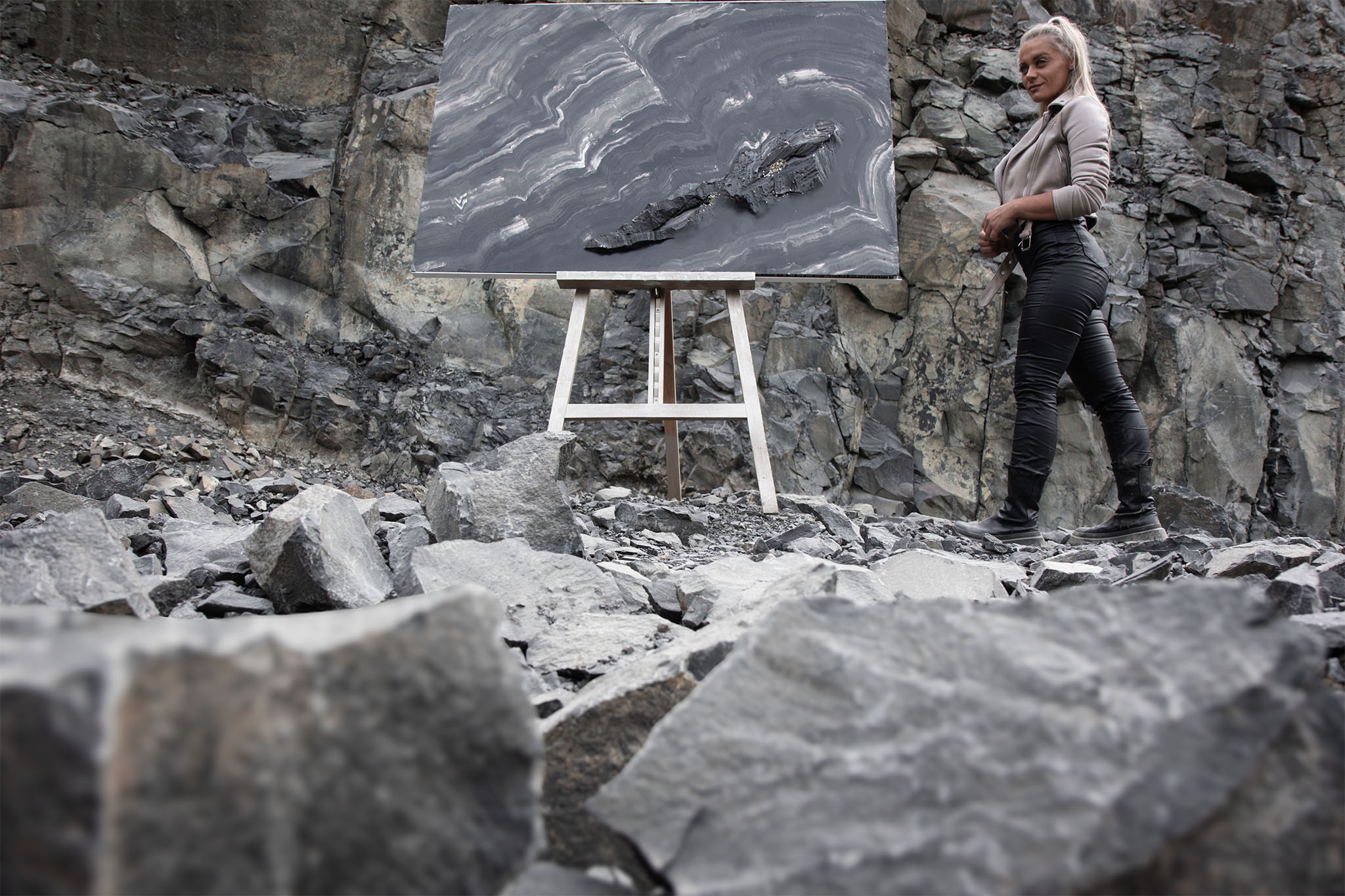 Die Künstlerin Jennit steht in einem Steinbruch seitlich neben einem großen, dunkelgrauen Kunstwerk von ihr, der Serie Marbleous.