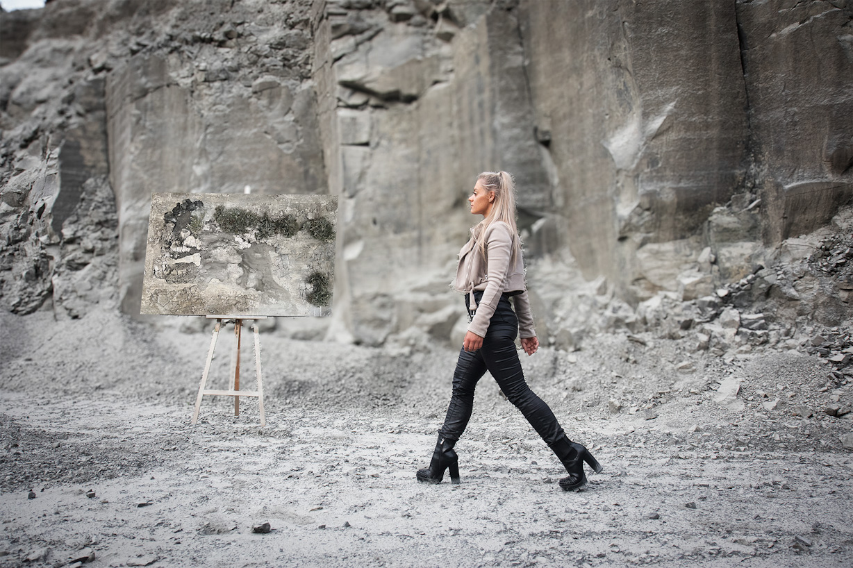 Die Künstlerin Jennit geht mit entschlossenem Schritt durchs Bild. Im Hintergrund sieht man einen Steinbruch und man sieht ein Kunstwerk mit Moos in taupe auf einer Staffelei